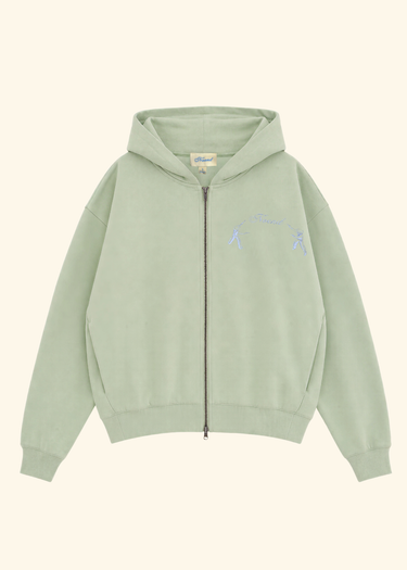 A sage green zip-up hoodie with a small light blue embroidered graphic on the left chest, displayed flat against a solid cream background.