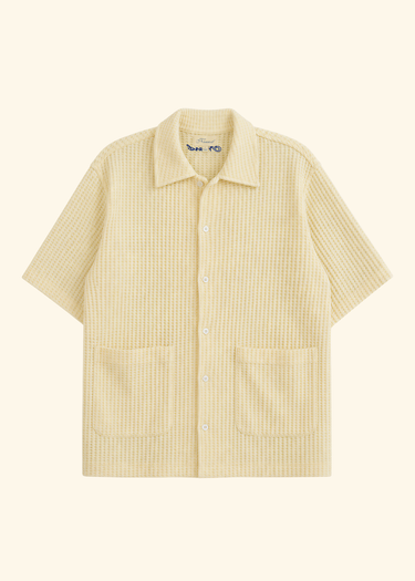 A short-sleeve button-up shirt in a light cream or pale yellow color with a fine textured grid pattern, featuring a pointed collar and two front patch pockets, shown against a solid cream background.