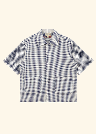 A short-sleeve button-up shirt with a small-scale black and white gingham pattern, featuring a pointed collar, white buttons, and two front patch pockets, displayed flat against a solid cream background.