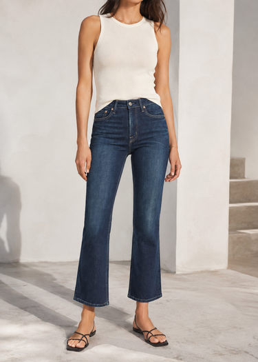 A model wearing dark blue cropped flare jeans, a white ribbed tank top, and black strappy flat sandals, standing in a bright, modern white interior setting.