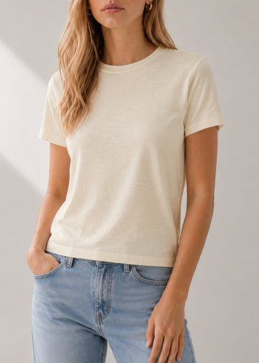 A woman wearing a short-sleeve crewneck t-shirt in a light cream color, paired with light blue jeans, shown from the neck down against a neutral studio background.