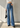 A model wearing high-waisted, wide-leg blue denim jeans with a tie waist, paired with a white long-sleeve crop top, black pointed shoes, and holding a black handbag against a textured gray wall.
