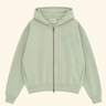 A sage green zip-up hoodie with a small light blue embroidered graphic on the left chest, displayed flat against a solid cream background.