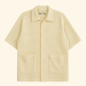 A short-sleeve button-up shirt in a light cream or pale yellow color with a fine textured grid pattern, featuring a pointed collar and two front patch pockets, shown against a solid cream background.