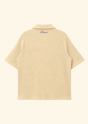 A short-sleeve button-up shirt with a fine gingham check pattern in beige and white, shown from the back on a solid cream background. A small blue script logo is visible on the inside neck label.