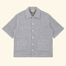 A short-sleeve button-up shirt with a small-scale black and white gingham pattern, featuring a pointed collar, white buttons, and two front patch pockets, displayed flat against a solid cream background.