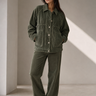 A woman stands in a minimalist studio wearing an olive green corduroy button-up jacket and matching wide-leg corduroy pants.