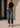 A woman wearing a black sleeveless top and dark gray high-waisted cropped jeans with rolled hems, standing outdoors against a concrete wall.