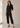 Woman wearing a black jumpsuit standing in a well-lit room with sunlight filtering through.