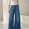 A model wearing a pair of high-waisted, wide-leg blue denim jeans with a beige knit top and white pointed-toe heels, standing in a minimalist, sunlit studio setting.