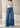 A model wearing a pair of high-waisted, wide-leg blue denim jeans with a beige knit top and white pointed-toe heels, standing in a minimalist, sunlit studio setting.