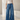 A model wearing a pair of high-waisted, wide-leg blue denim jeans with a beige knit top and white pointed-toe heels, standing in a minimalist, sunlit studio setting.