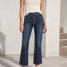 A model wearing dark blue cropped flare jeans, a white ribbed tank top, and black strappy flat sandals, standing in a bright, modern white interior setting.