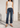 A model wearing dark blue cropped flare jeans, a white ribbed tank top, and black strappy flat sandals, standing in a bright, modern white interior setting.