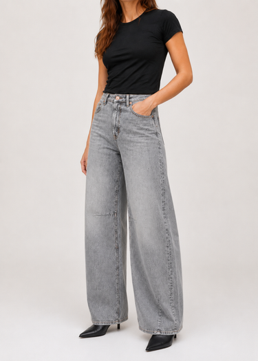 A model wearing light gray wide-leg denim jeans, a black short-sleeve t-shirt, and black pointed-toe heels, standing against a plain light gray studio background.