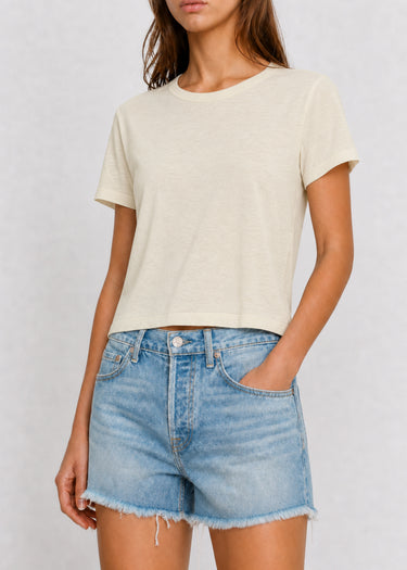 A woman wearing a light cream-colored short-sleeve crop top and light-wash blue denim shorts, standing against a neutral light gray background.