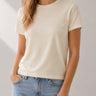 A woman wearing a short-sleeve crewneck t-shirt in a light cream color, paired with light blue jeans, shown from the neck down against a neutral studio background.