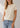 A woman wearing a short-sleeve crewneck t-shirt in a light cream color, paired with light blue jeans, shown from the neck down against a neutral studio background.