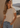 A woman wearing a light beige short-sleeve crewneck t-shirt and light blue jeans, leaning against a textured stucco wall with a scenic coastal view in the background.