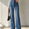 A model wearing high-waisted, wide-leg blue denim jeans with a tie waist, paired with a white long-sleeve crop top, black pointed shoes, and holding a black handbag against a textured gray wall.