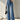 A model wearing high-waisted, wide-leg blue denim jeans with a tie waist, paired with a white long-sleeve crop top, black pointed shoes, and holding a black handbag against a textured gray wall.