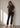 Woman wearing a black t-shirt and black jeans standing against a light wall with sunlight casting shadows.